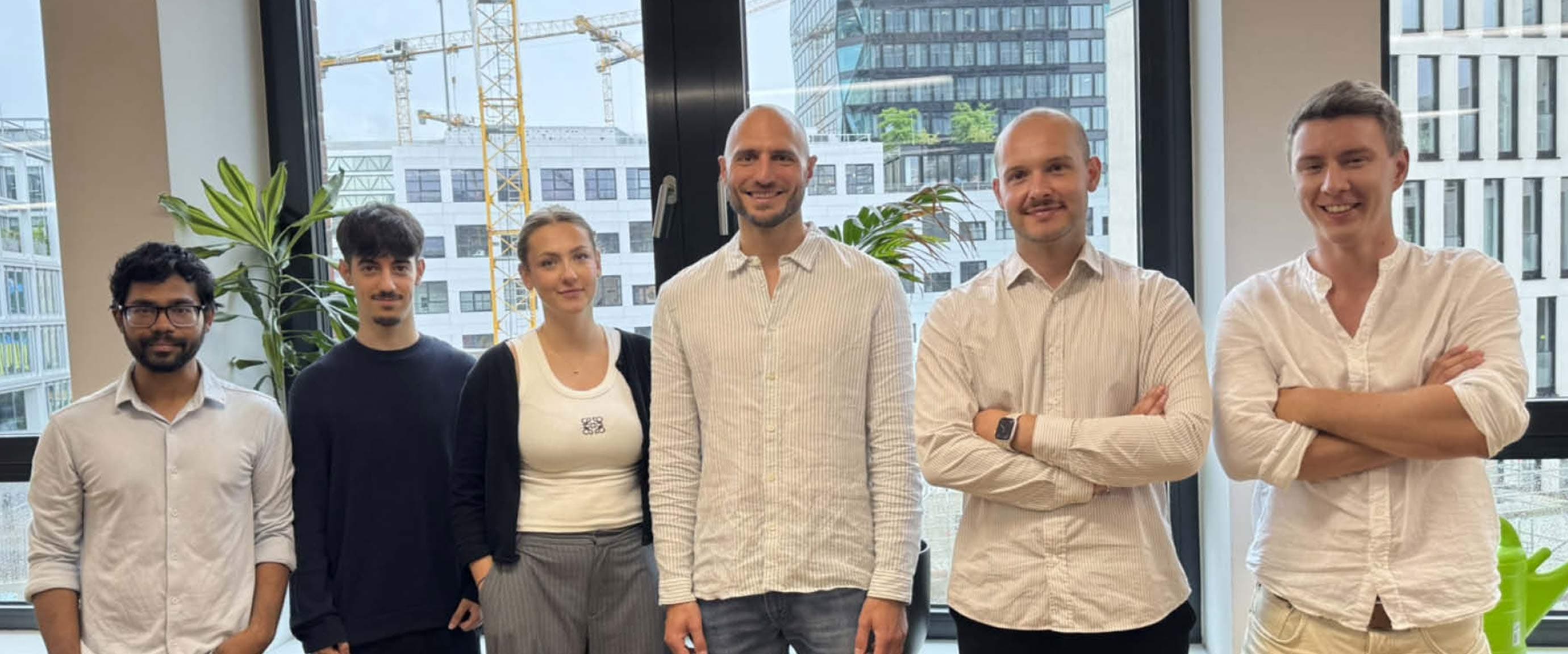 Group photo of the Complioty team in a modern office in front of large windows overlooking office buildings. Six team members are standing side by side.