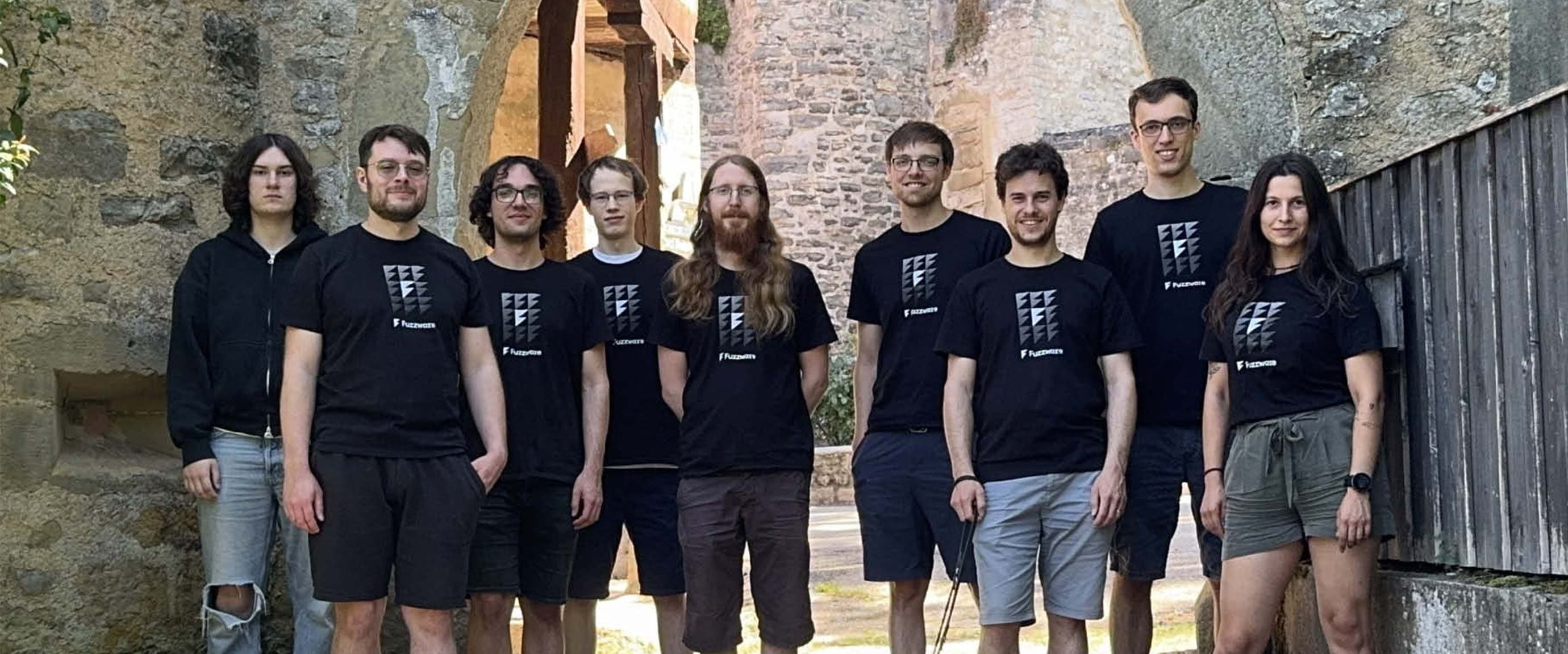 Group photo of the Fuzzware team outdoors in front of historic stone walls. Several team members wear black company shirts, with a dog lying in the foreground.